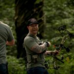 Two men smile at each other in the woods while holding archery equipment