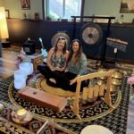 Two smiling women sit on a floor surrounded by bowls, and musical instruments