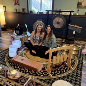 Two smiling women sit on a floor surrounded by bowls, and musical instruments