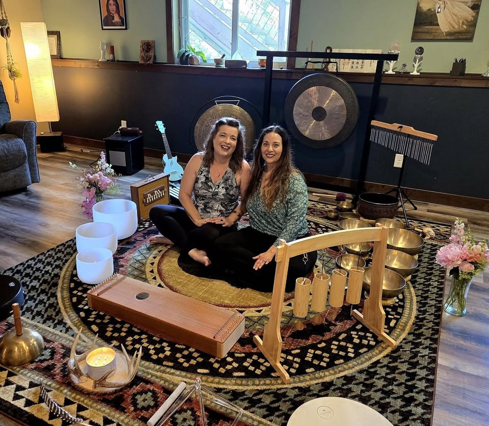 Two smiling women sit on a floor surrounded by bowls, and musical instruments