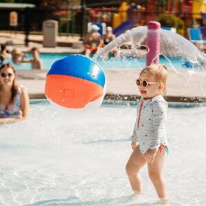 A toddler in sunglasses stands in a shallow pool, smiling as a colorful beach ball floats nearby.
