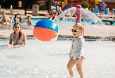 A toddler in sunglasses stands in a shallow pool, smiling as a colorful beach ball floats nearby.