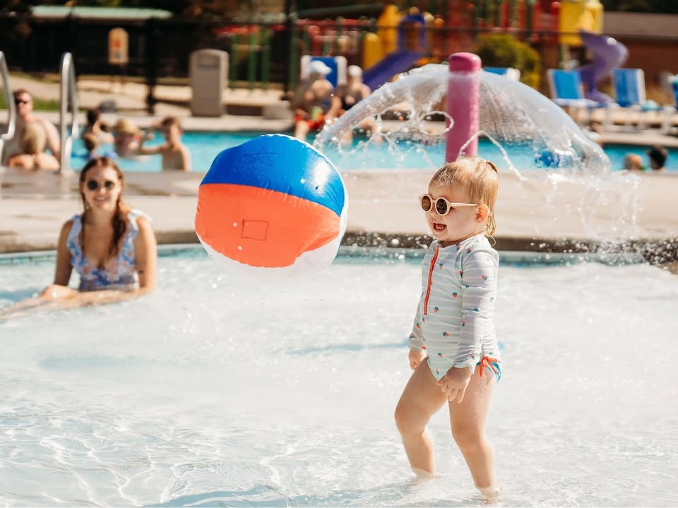 A toddler in sunglasses stands in a shallow pool, smiling as a colorful beach ball floats nearby.